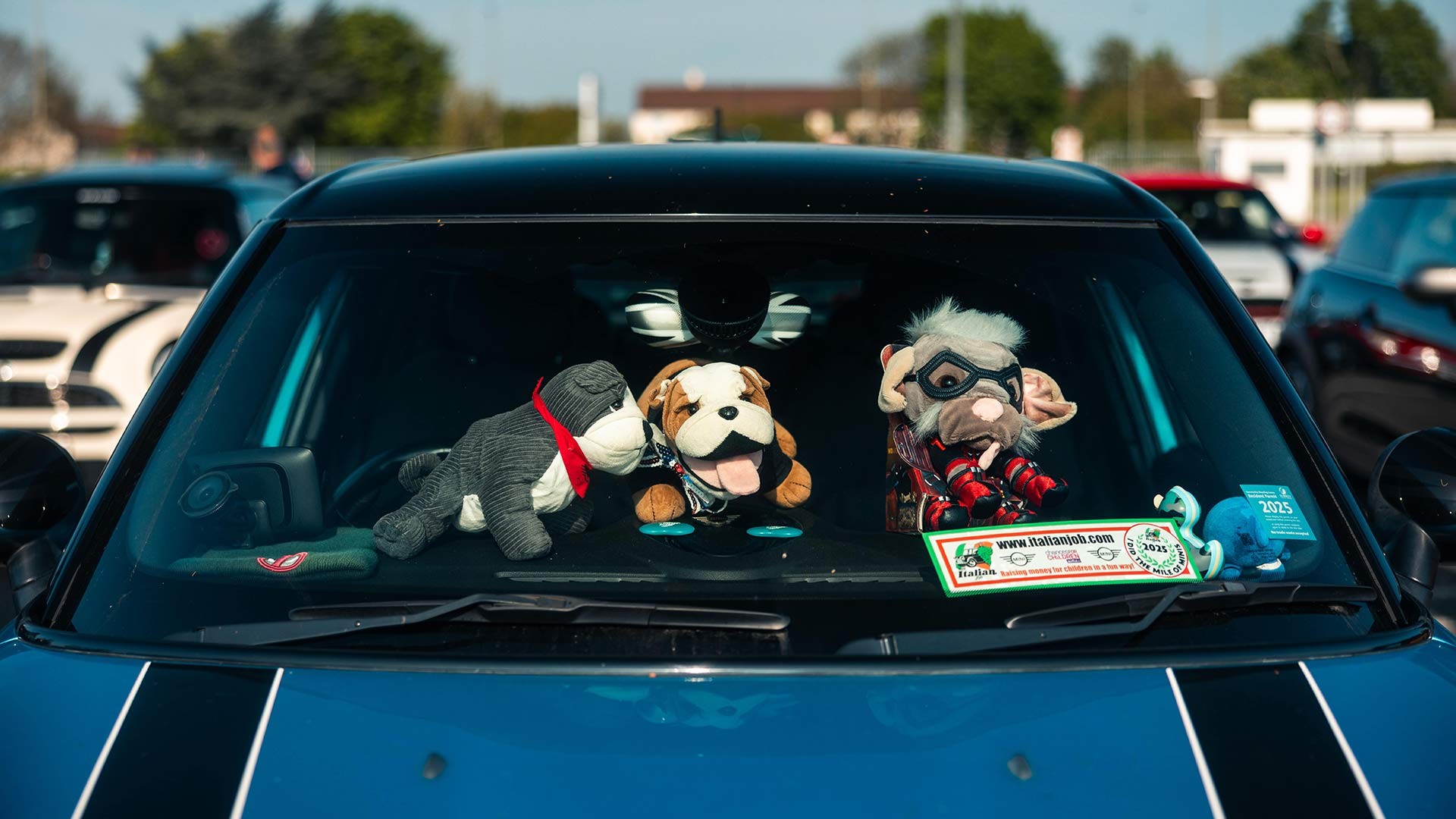 A car dashboard displays three plush toys: a dog, a bulldog, and a whimsical character with goggles. 